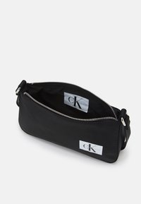 Czarna nylonowa kopertówka o gładkiej fakturze, z srebrnym zamkiem błyskawicznym i metką z logo wewnątrz. Kompaktowy prostokątny kształt idealny na niezbędne przedmioty.