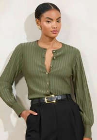 Olivgrüner gerippter Cardigan mit Rundhalsausschnitt, goldenen Knopfverschlüssen und langen Ärmeln, kombiniert mit hoch-taillierten schwarzen Hosen und einem schwarzen Gürtel.