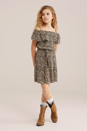 Jong meisje met lang krullend haar, gekleed in een off-shoulder jurk met luipaardprint, witte sokken en bruine enkellaarsjes, staand met gekruiste benen.