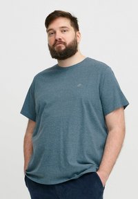 Teal színű, kerek nyakú póló puha, texturált anyagból. Rövid ujjú, és egy kisebb logó található a mellrészen. A modell a zsebeibe dugott kezekkel áll.