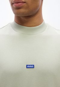 Κοντινό πλάνο ενός ατόμου που φοράει ανοιχτοπράσινο φούτερ με ένα μικρό μπλε ορθογώνιο ταμπελάκι με το λογότυπο "HUGO" στο στήθος.