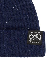 Bonnet tricoté bleu marine avec une texture côtelée, ornée de petites touches multicolores et d'une étiquette noire affichant un motif de montagne et de soleil.