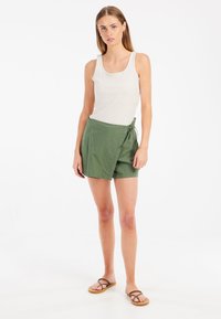 Groene wikkelshorts van lichtgewicht stof, met een striksluiting en een relaxte pasvorm. Gecombineerd met een witte geribbelde tanktop en teenslippers.