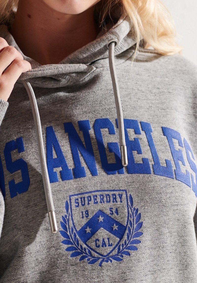 Gros plan d'une personne portant un sweat à capuche gris avec le texte bleu « LOS ANGELES » et un emblème bleu avec le texte « SUPERDRY 1954 CAL ».