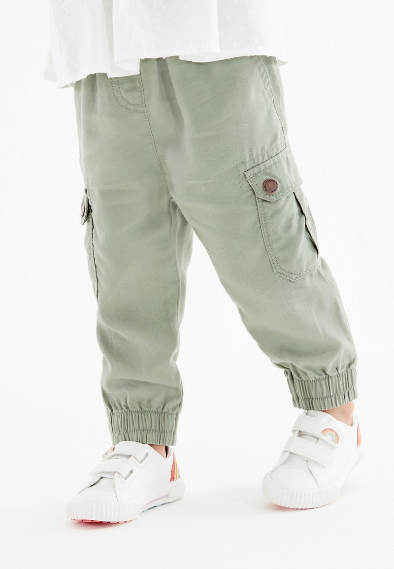 Enfant portant un pantalon cargo vert clair avec des poignets élastiques et des baskets blanches avec des détails arc-en-ciel sur les côtés.