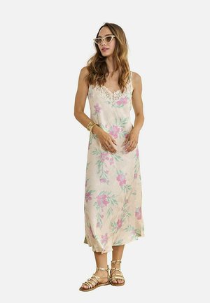 Mujer con un vestido midi sin mangas color crema con estampado floral rosa, escote de encaje, brazaletes dorados, sandalias con tiras y gafas de sol estilo cat-eye.