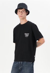 Black cotton t-shirt with short sleeves, featuring white script logo on the left chest. Worn with denim jeans and a black bucket hat.