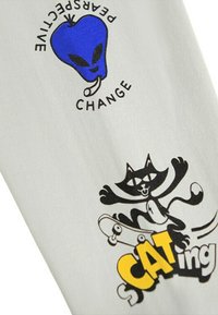 Bílá tkanina s dvěma tištěnými motivy: modré jablečné jablko ve tvaru mimozemšťana s textem "Změna perspektivy" a černá kreslená kočka na skateboardu s nápisem "SCATing."