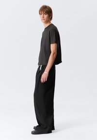 Giovane con i capelli corti castani, che indossa una maglietta nera larga, pantaloni neri larghi con coulisse bianca e scarpe nere, in posizione di profilo.