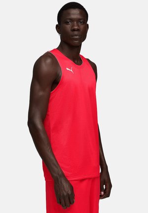 Atletyczny ciemnoskóry mężczyzna stojący, ubrany w czerwoną bezrękawnikową sportową bluzkę Puma i pasujące krótkie spodenki na jednolitym jasnym tle.