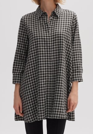 Vrouw die een lang zwart-wit geruit overhemd met driekwartmouwen draagt over een zwarte broek, staand met haar armen ontspannen langs haar zij.