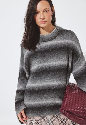 Moteris su ilgais tamsiais plaukais, vilkinti pilką dryžuotą megztinį, nešanti didelę austą tamsiai rožinę rankinę virš pledinės sijono.