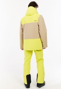 Geel en beige gekleurde blokjasje met een capuchon, gecombineerd met felgele sneeuwbroek en zwarte accenten op de zoom en laarzen.