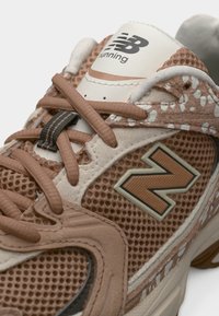 Κοντινό πλάνο καφέ και μπεζ παπουτσιού τρεξίματος New Balance με ύφασμα δίχτυ, διακοσμητικά μοτίβα και ορατά λογότυπα New Balance στη γλώσσα και στην πλευρά.