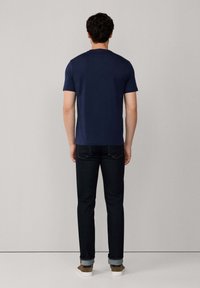 Hombre con camiseta azul marino, pantalones vaqueros oscuros remangados y zapatos marrones, de pie frente a una pared lisa de color gris claro.