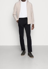 Camicia a maniche lunghe beige chiaro sopra una t-shirt bianca a girocollo, pantaloni neri attillati e scarpe grigio scuro con suole chiare, in piedi su un pavimento grigio.