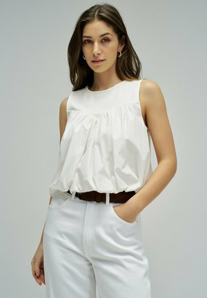 Jeune femme aux longs cheveux bruns portant un haut blanc sans manches à volants, un pantalon blanc et une ceinture marron foncé, posant avec une main dans la poche.