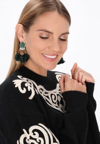 Orecchini con pietre preziose verdi e frange con accenti argentati. Maglione nero con ricami ornamentali color crema. Tessuto morbido; vestibilità rilassata.