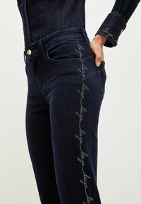 Tummat farkut, joissa on monimutkaisia hopeakoristeita sivusauman varrella, muotoiltu malli ja klassiset taskudetails.