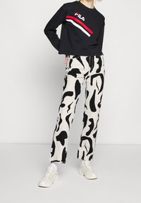 Personne portant un sweat FILA noir, un pantalon blanc avec un motif abstrait noir, et des baskets blanches épaisses, debout devant un mur uni.