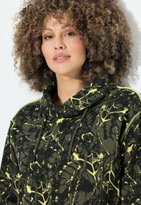 Sudadera negra y verde con un patrón amarillo veteado, que presenta un cuello alto, cordones y una tela texturizada.