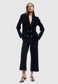 Czarna dwurzędowa marynarka z złotymi guzikami, połączona z szerokimi spodniami. Modelka nosi czarne sandały na obcasie. Gładki, dopasowany materiał.