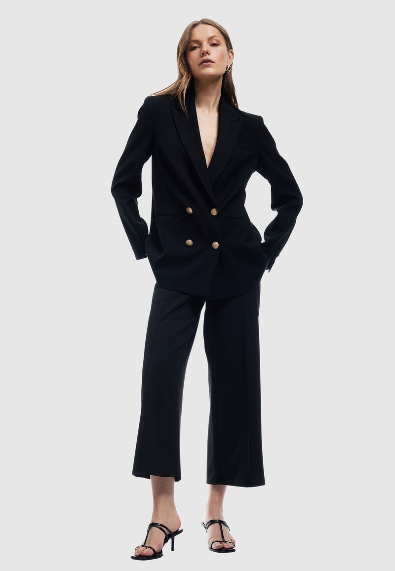 Czarna dwurzędowa marynarka z złotymi guzikami, połączona z szerokimi spodniami. Modelka nosi czarne sandały na obcasie. Gładki, dopasowany materiał.
