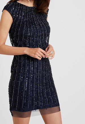 Kvinde iført en ærmeløs sort kjole med lodrette palietstriber og gennemsigtig kant, hænder foldet foran, ensartet lysegrå baggrund.