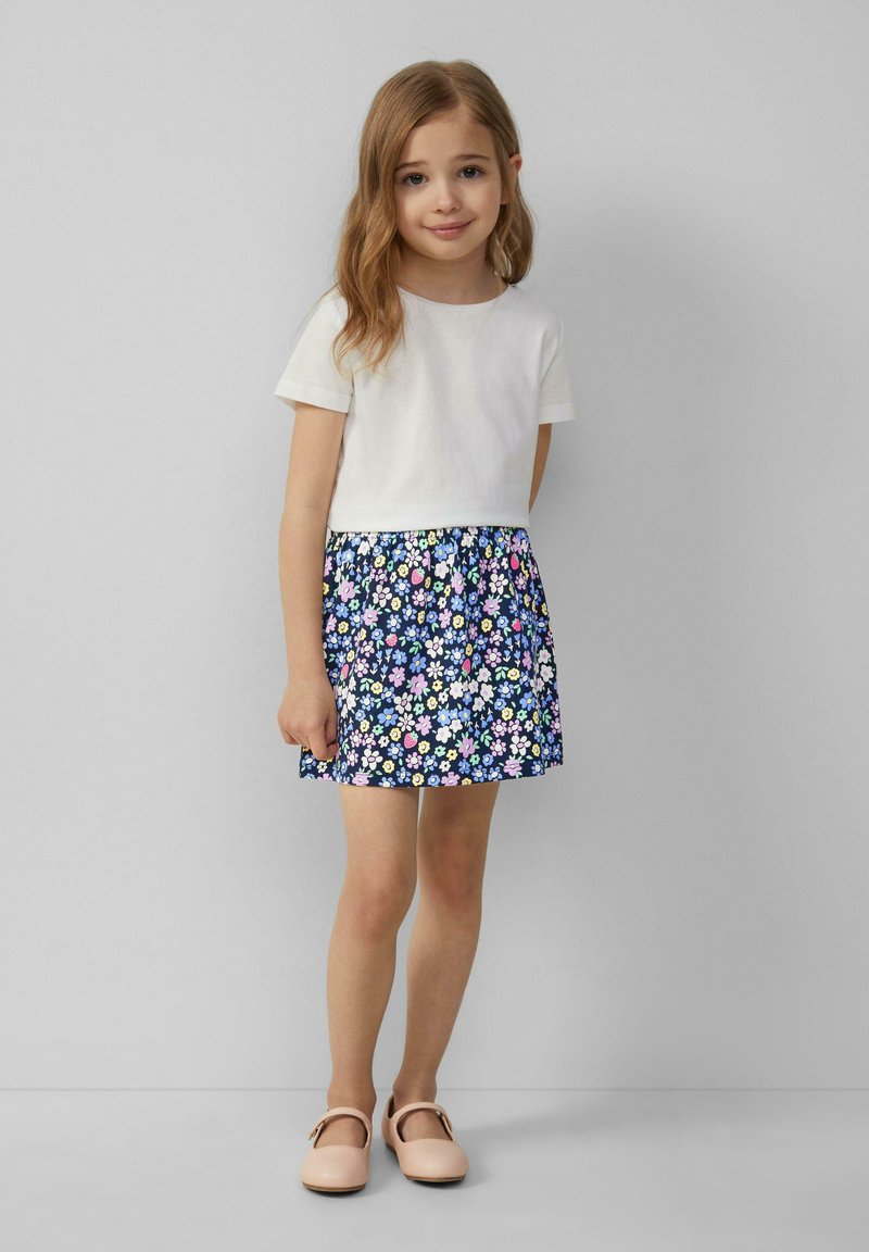 Witte kortmouwtop gecombineerd met een blauwe bloemenrok. De rok heeft een verscheidenheid aan kleurrijke bloemen in een geplooid ontwerp. Lichte schoenen.