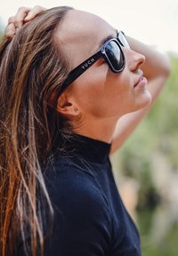 Černé sluneční brýle s logem "VUCH" na boku, nošené osobou s dlouhými hnědými vlasy, v úhlovém bočním pohledu, oblečenou v černém topu s vysokým límcem.