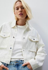 Hvid denimjakke med metalnagledekorationer, to forlommer og trykknapper, båret over en hvid ribbet skjorte og blå jeans.