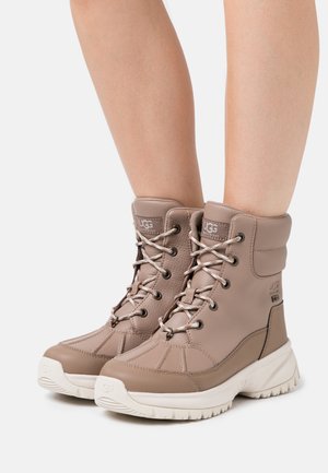 Bottes en cuir fauve imperméables avec cheville rembourrée, surface texturée et semelle crème contrastante. Dotées d'œillets métalliques et de lacets décoratifs.