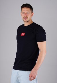 Mann mit kurzen Haaren und Bart, der ein schwarzes T-Shirt mit rotem Logo und hellblauen Jeans trägt und vor einem grauen Hintergrund steht.