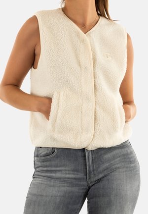 Femme portant un gilet en polaire crème sans manches avec des poches et un jean gris, les mains dans les poches, sur fond blanc.