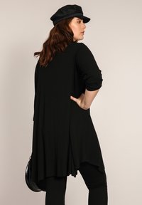 Un haut long noir à côtes avec un ourlet asymétrique fluide, associé à des leggings noirs et une casquette noire. La texture est douce, au design décontracté.