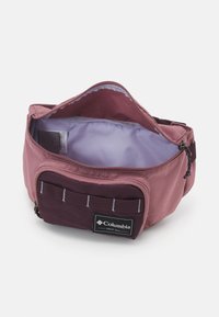 Aukikirjaimella sinettinä varustettu avonainen kaksivärinen purppura vyölaukku, jossa on näkyvä vaalea sisävuori ja etutasku, johon on painettu Columbia-logo.