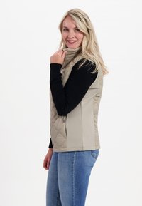 Beige gesteppte Weste mit hohem Kragen, kombiniert mit einem schwarzen Langarmshirt. Die Weste hat einen figurbetonten Schnitt und Reißverschlusstaschen.