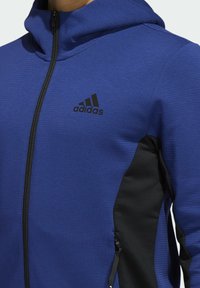 Sweat à capuche zippé bleu avec panneaux latéraux noirs, tissu texturé, col montant et logo Adidas sur la poitrine. Design léger.