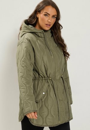 Femme portant une veste matelassée à capuche de couleur vert olive avec taille à cordon et poches à rabat, debout devant un fond clair uni.