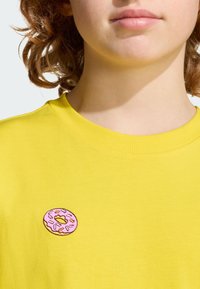 Camiseta amarilla de manga corta con cuello redondo y un pequeño bordado de donut rosa en el pecho, con coloridos sprinkles.