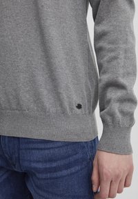 Grå strikkegenser med ribbestrikket kant og mansjetter. Har en liten svart logo på den nedre siden. Kombinert med blå denim jeans.
