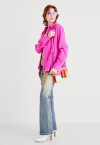 Bright pink button-up shirt, beige belt, faded blue flared jeans, white heeled boots, and a multicolored small handbag with graphic accents.