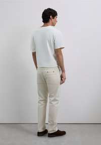 Hombre vestido con una camiseta color blanco roto y pantalones beige, de pie mirando hacia atrás frente a un fondo gris claro liso, con zapatos marrones.
