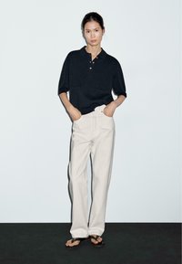 Polo shirt blu scuro a maniche corte con due tasche frontali, abbinato a pantaloni beige chiaro larghi e ciabatte nere su uno sfondo neutro.