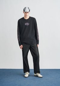 Juoda ilgomis rankovėmis marškinėliai su centriniu grafikos elementu ir violetiniais akcentais, derinami su tamsiomis kelnėmis ir baltais sportbačiais. Modelis dėvi dryžuotą kepuraitę.