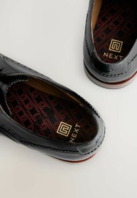 Zwarte gepatineerde leren loafers met een contrasterende bruine zool; interieur met een patrooninlegzool met een gouden logo. Gladde textuur, klassiek ontwerp.