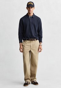 Marineblaues Poloshirt, beige Chinos und dunkle Sneakers. Das Outfit beinhaltet eine marineblaue Mütze mit gelbem Text und einen braunen Gürtel.