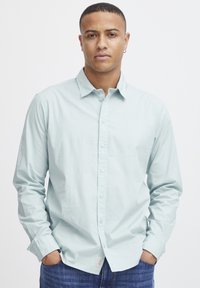 Camisa de botones azul claro hecha de algodón, con cuello puntiagudo, mangas largas y un corte regular. Llevada sobre unos vaqueros azules.