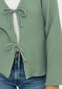 Top envolvente texturizado verde claro con escote en V y cierres de lazo; combinado con jeans de mezclilla azul que presentan un botón visible y un bolsillo.