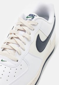 Weißer Sneaker mit navy- und grünem Nike-Logo, strukturiertem Obermaterial, beigen Schnürsenkeln und gummierter Sohle. Verfügt über perforierte Details an der Zehe.